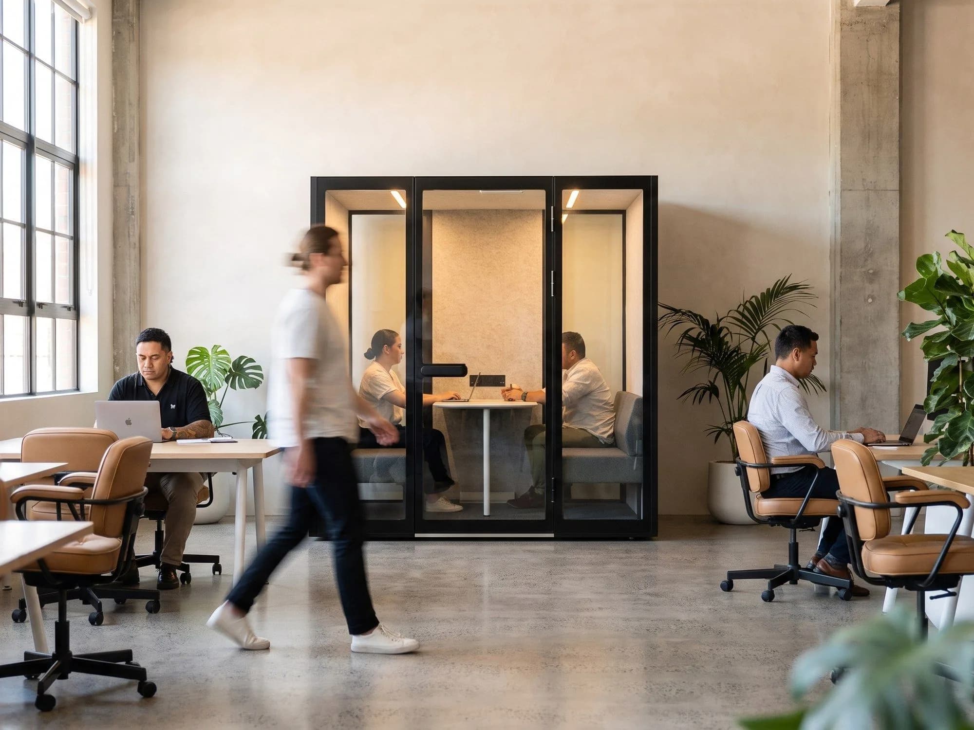 Two people meeting inside a Peacepod in an open-plan office, colleagues working at desks alongside