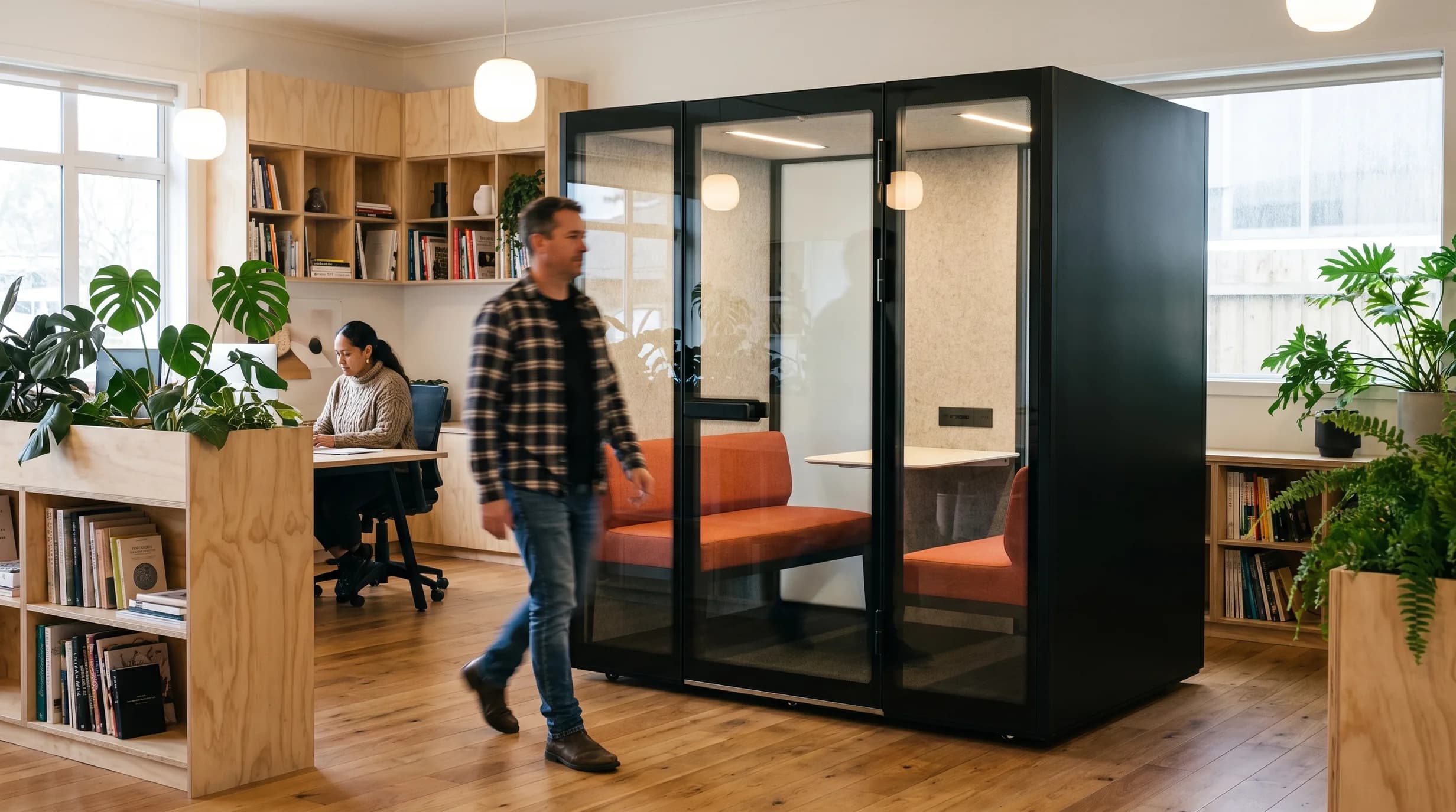 L4-Pod meeting pod in a warm plant-filled timber office, a person walking past; coral seating visible through the glazed doors