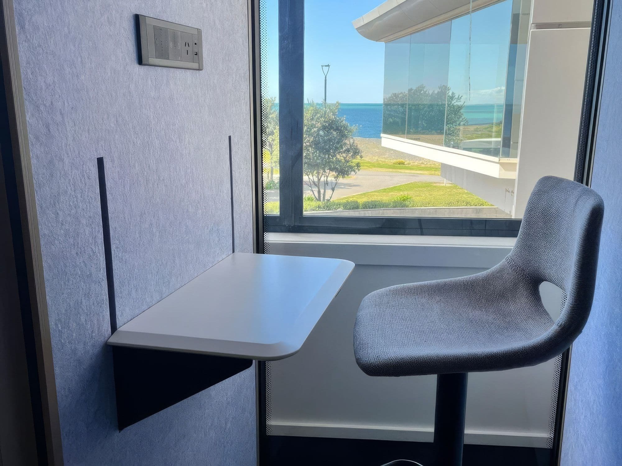 Inside an S-Pod at a client site, with the white sit-stand desk lowered beside a grey height-adjustable stool, and a window looking out to the sea