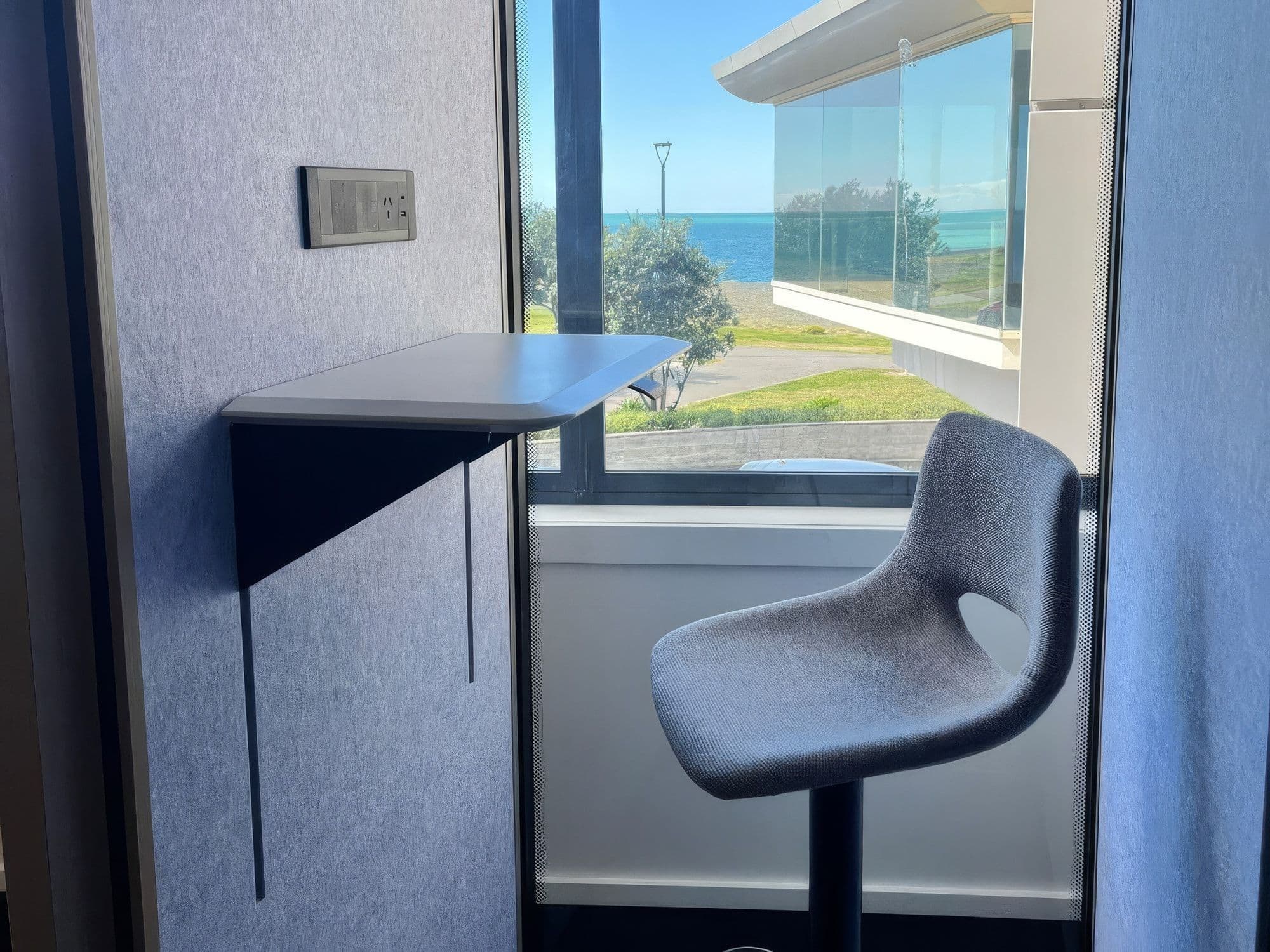 Inside an S-Pod at a client site, with the white sit-stand desk in its raised position beside a grey height-adjustable stool, and a window looking out to the sea